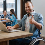 Homme souriant dans un fauteuil roulant manuel donnant un coup de pouce assis à une table à l'aide d'un ordinateur portable.