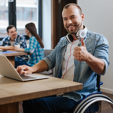 Smiling man in a manual wheelchair giving a thumbs up sitting at a table using a laptop